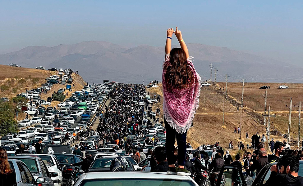 En kvinna står på ett biltak med ryggen mot kameran. Massor av människor köar och demonstrerar på en väg i Iran.