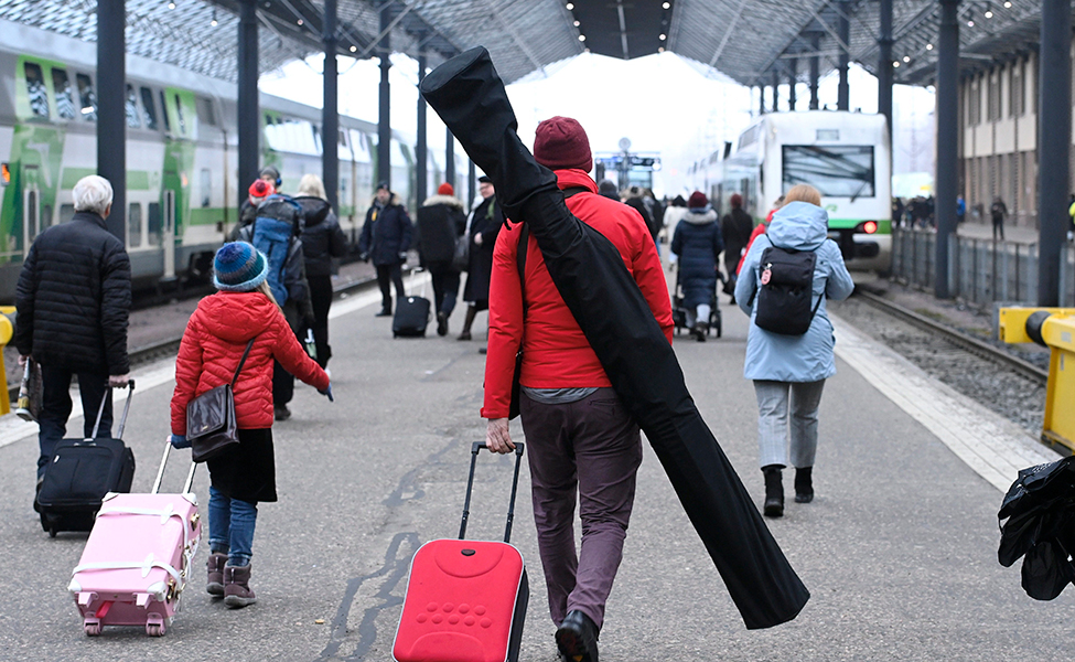 Bild från en järnvägsstatione. Man ser människor bakifrån. Många drar resväskor, en har skidor.
