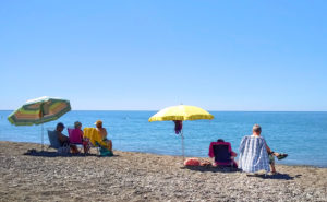Personer sitter under parasoll på en havsstrand. Solen skiner.