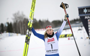 En ung kvinna med skidor i den ena handen och stavar i den andra. Hon ser glad ut och höjer båda armarna.
