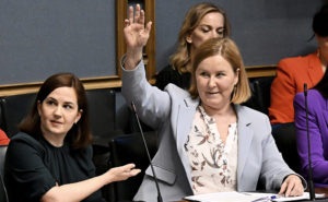 Ministrarna Sanni Grahn-Laasonen och Kaisa Juuso i riksdagens plenisal. Minister Kaisa Juuso räcker upp handen.