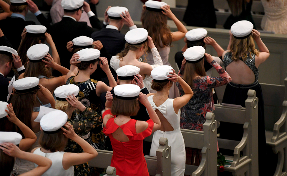 På bilden finns många studenter i festkläder. De står i rader. Alla studenterna sätter studentmössan på sitt huvud. Bilden visar studenterna bakifrån.