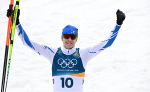 En man som ser glad ut och lyfter upp sina händer i en segergest. Han är klädd i skidåkarkläder och håller ett par skidor i ena handen.